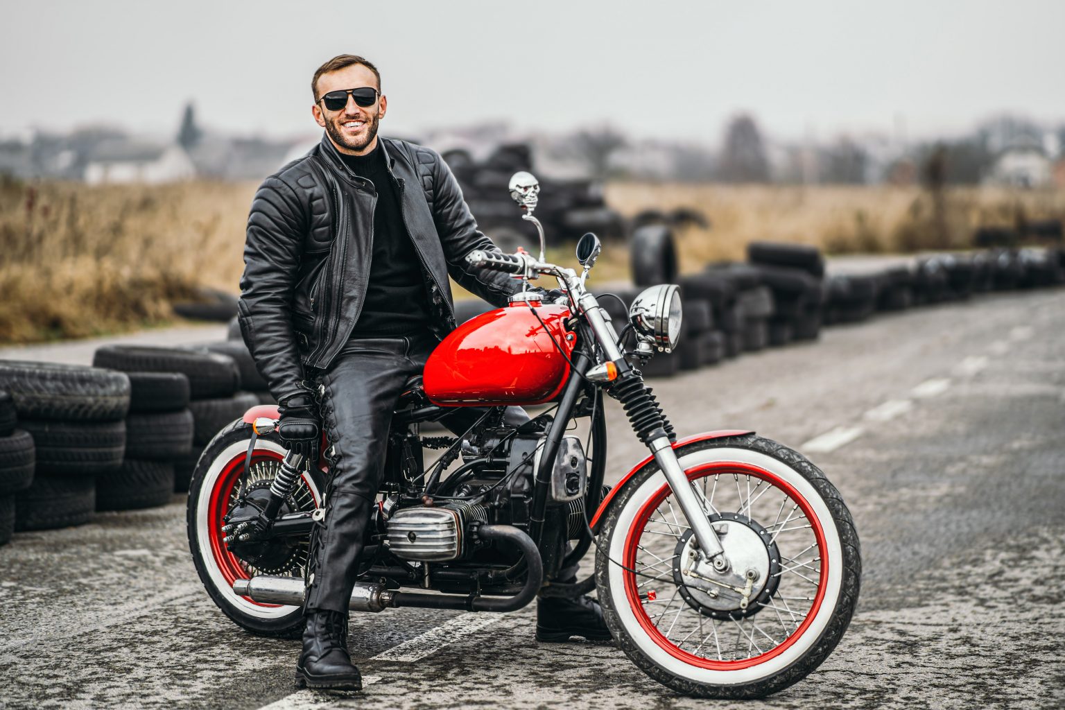 Red motorbike with rider. A man in a black leather jacket and pants stands sideways in the middle of the road. Tires are laid on the background.